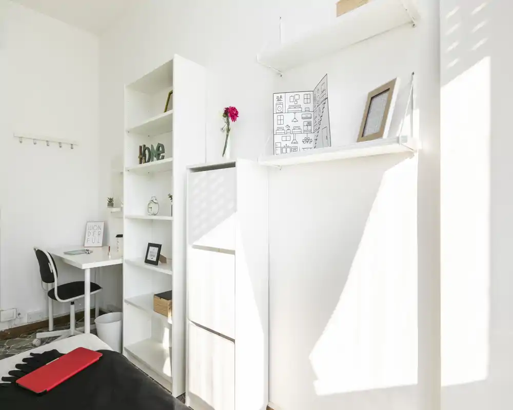 Chambre lumineuse et minimaliste avec lit au premier plan, étagères blanches et petit bureau contre le mur ; la lumière naturelle donne une atmosphère propre et aérée.