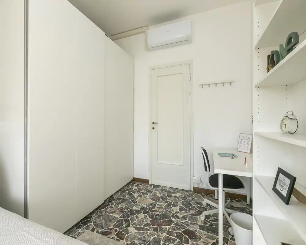 Petit coin chambre propre avec placard, bureau et étagères ; une partie du lit est visible sur le bord gauche. Tons neutres et sol en pierre à motifs donnent un espace minimaliste et bien éclairé, adapté à une personne seule.