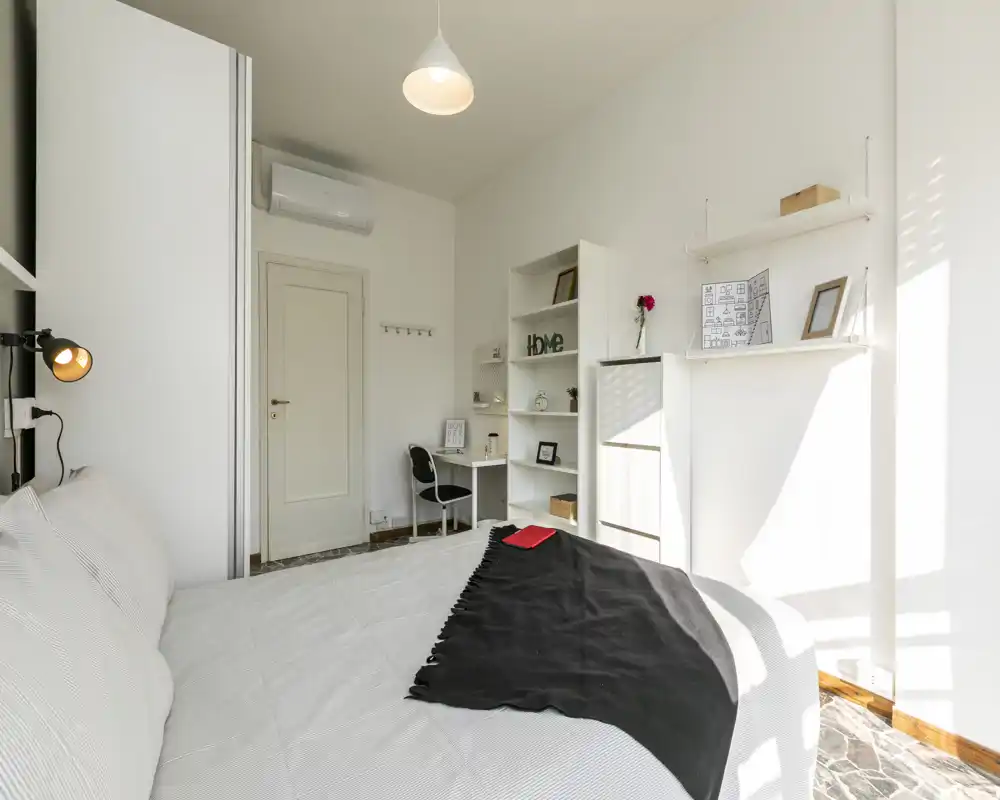 Chambre moderne et lumineuse avec un grand lit au premier plan, étagères minimalistes et un petit bureau près de la porte. La lumière naturelle inonde la pièce et la décoration neutre apporte une atmosphère apaisante et accueillante.