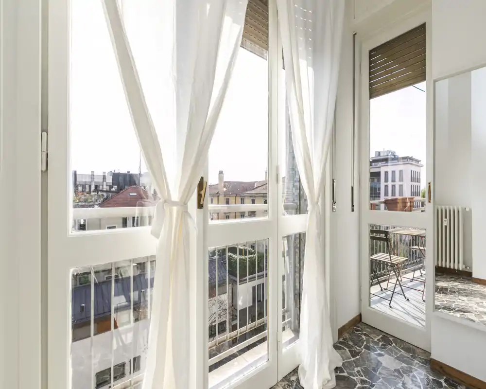 Vue lumineuse sur le balcon à travers de grandes fenêtres-fenêtres avec voilages, petite table et chaises extérieures et vue urbaine dégagée — idéale pour la lumière du matin.