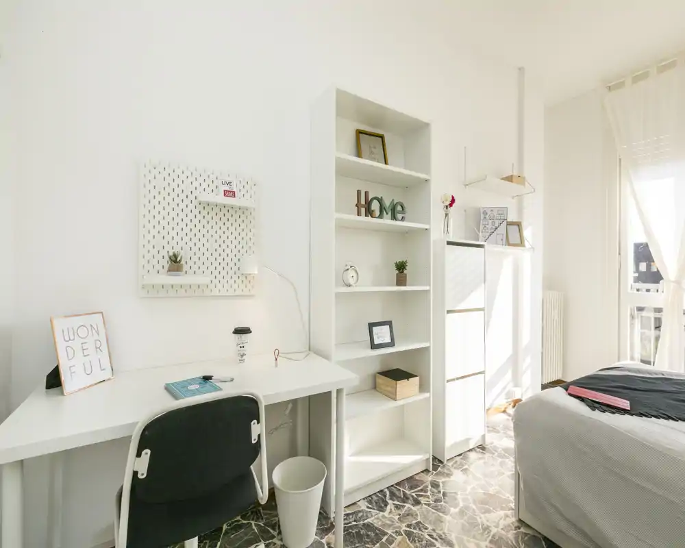 Chambre lumineuse et minimaliste avec lit bien fait près d'une fenêtre, bureau et étagères ouvertes — idéale pour une location étudiante ou individuelle.