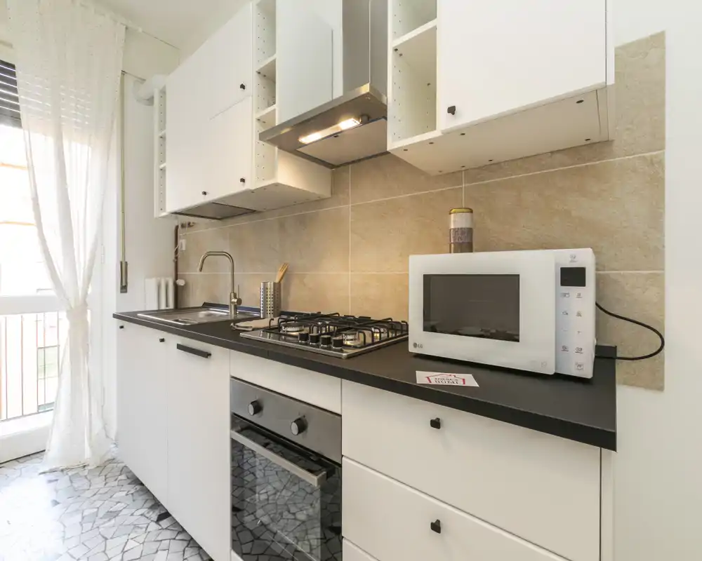 Cuisine moderne et lumineuse en style couloir avec plaque gaz, four encastré et micro-ondes sur plan de travail noir; carrelage clair et rideau léger pour une ambiance aérée.