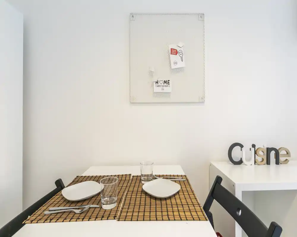 Coin repas minimaliste avec une petite table dressée pour deux, sets de table simples, assiettes et verres sur un mur blanc lumineux.
