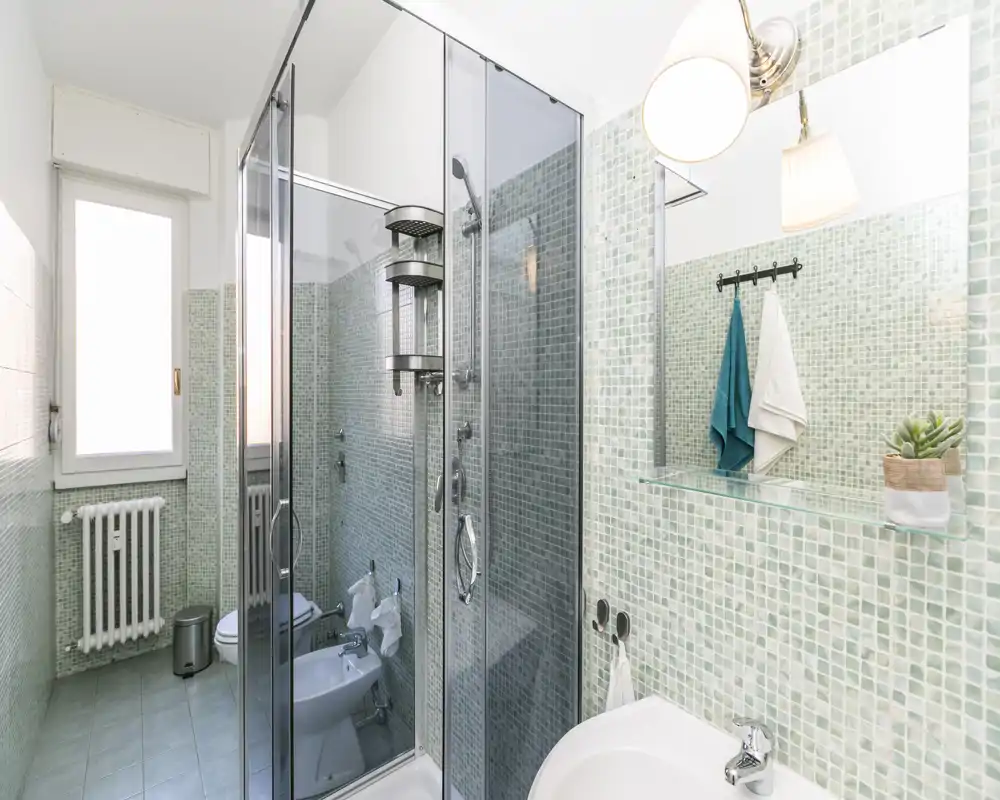 Salle de bains moderne et lumineuse avec douche vitrée, bidet et lavabo sur carreaux mosaïque vert pâle et grand miroir.