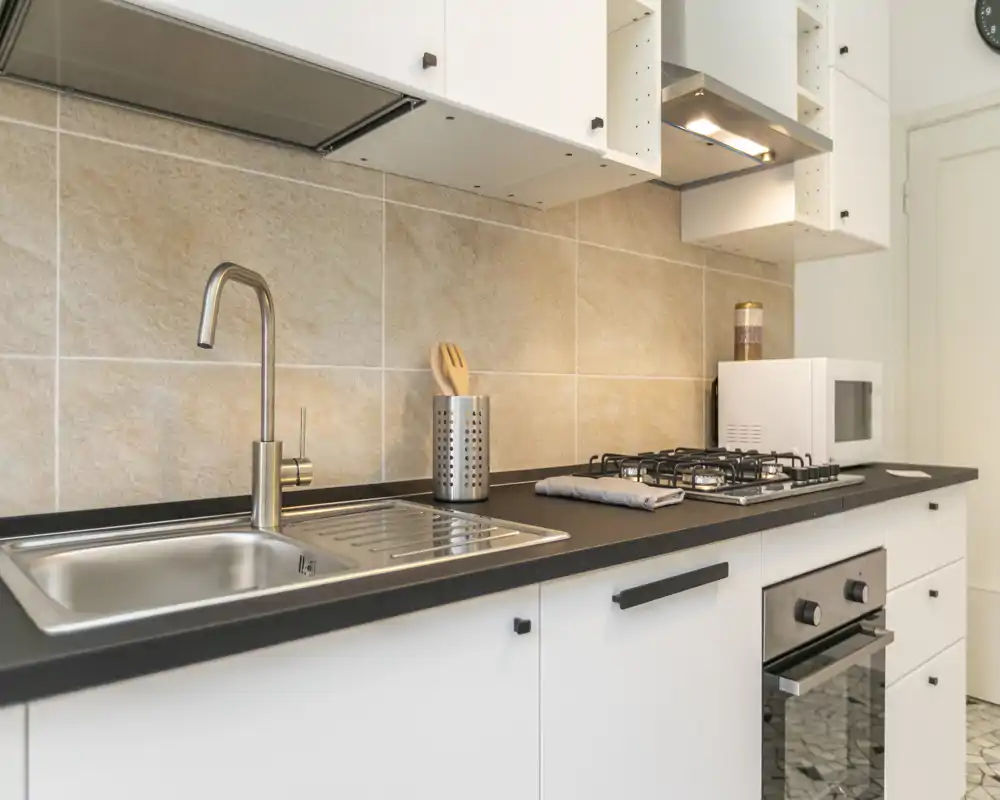 Cuisine moderne et lumineuse avec évier en acier inoxydable, plaque à gaz, four et micro-ondes ; crédence en carrelage clair et plan de travail soigné pour un espace de cuisson fonctionnel.
