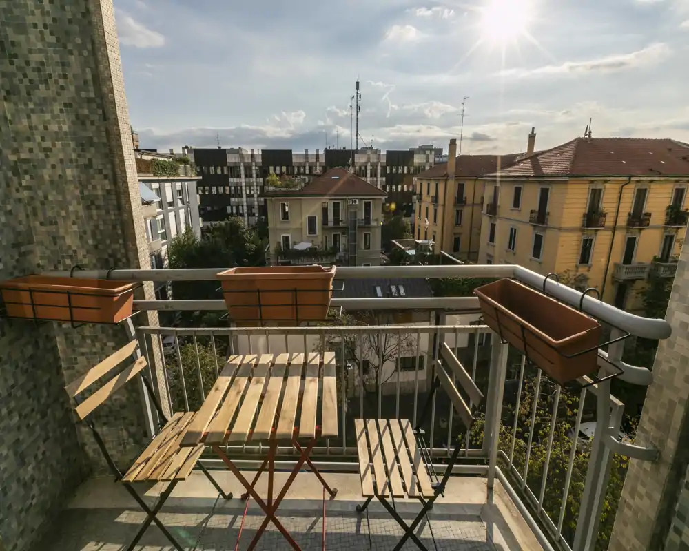 Balcon ensoleillé en ville avec deux chaises pliantes en bois et une petite table assortie, jardinières sur la rambarde et vue sur les immeubles voisins.