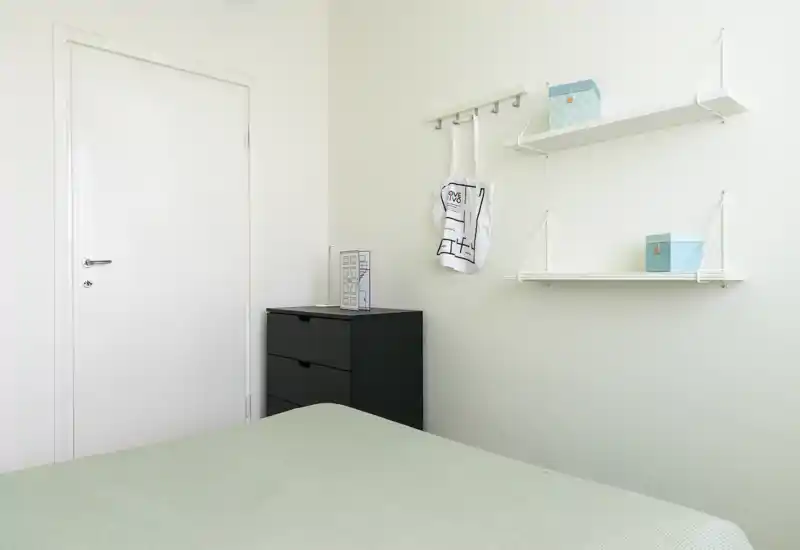 Camera da letto minimale e luminosa con letto in primo piano, un comodino nero e mensole bianche a parete con piccoli contenitori decorativi.