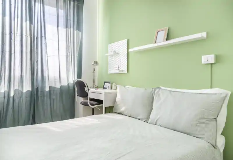 Camera da letto luminosa e ben curata con letto matrimoniale, pareti verde tenue e una scrivania accanto alla finestra che offre luce naturale.