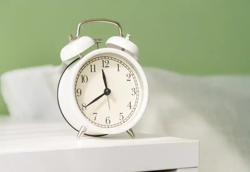 Primo piano di una sveglia da comodino su un tavolino con parte del letto in sfocatura; composizione luminosa, ordinata e accogliente.