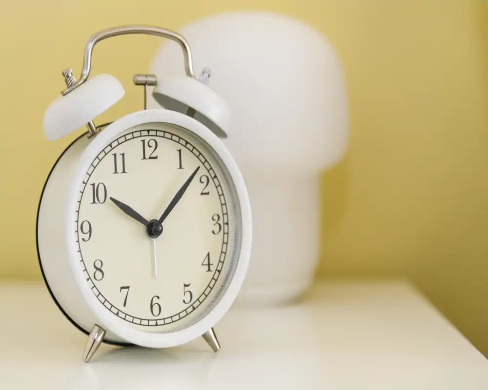 Primo piano di una sveglia classica sul comodino bianco con una lampada sfocata e toni caldi, perfetto per trasmettere un'atmosfera accogliente in camera da letto.