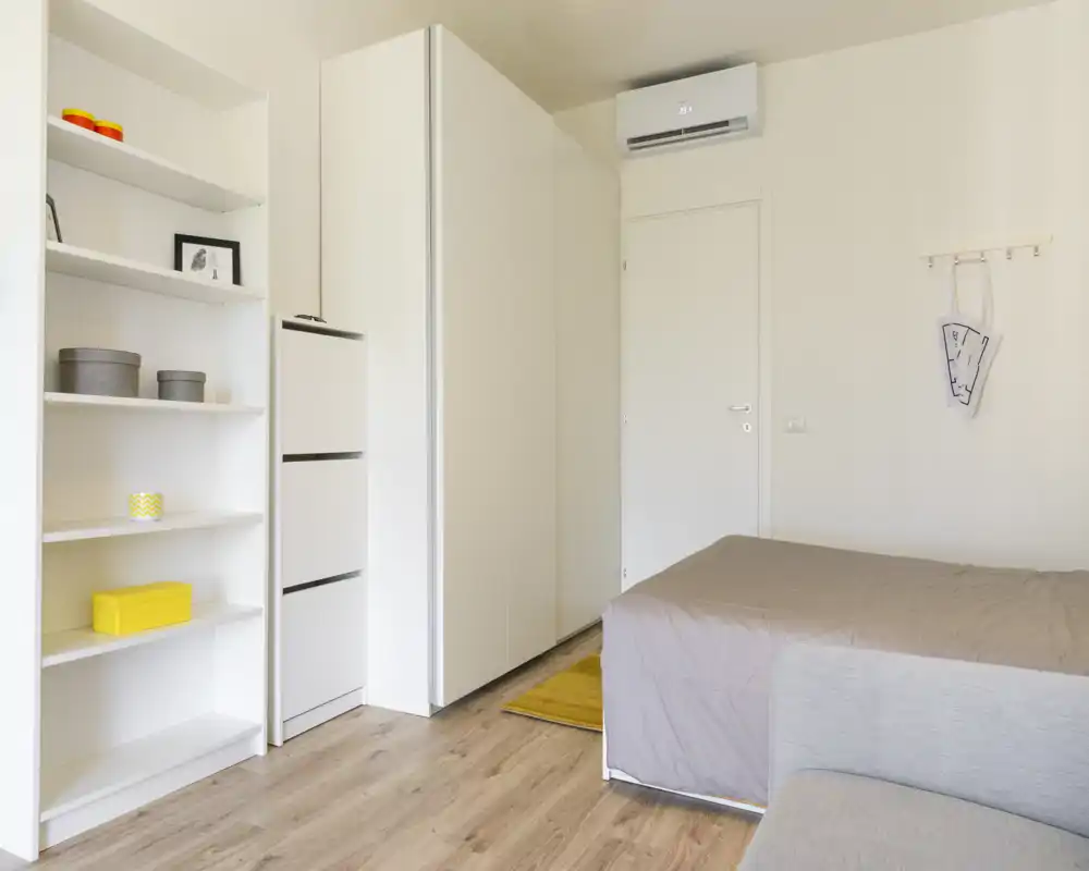 Camera da letto moderna e luminosa con letto matrimoniale parzialmente visibile, armadi bianchi e scaffalatura aperta con decor minimalista — pavimento in legno che dona calore all’ambiente.