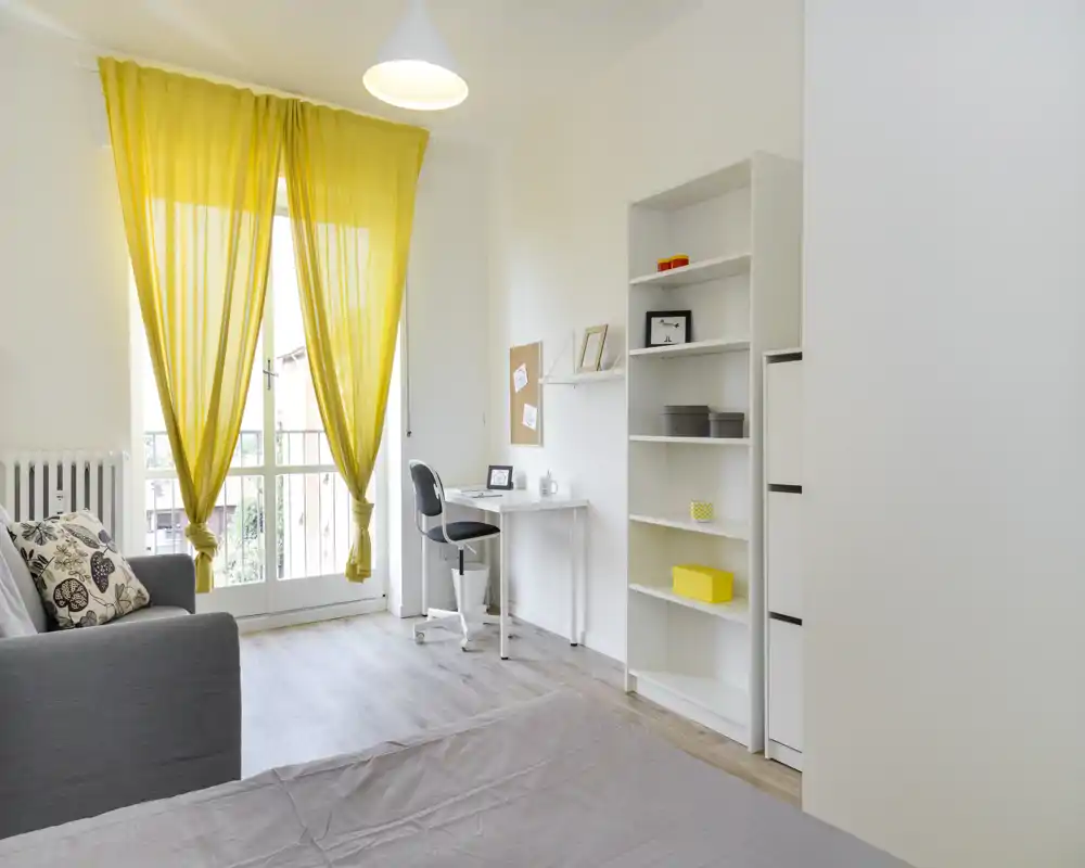 Camera da letto luminosa e moderna con letto in primo piano, una scrivania vicino alla grande finestra con tende gialle e scaffalatura aperta — elegante e ariosa.