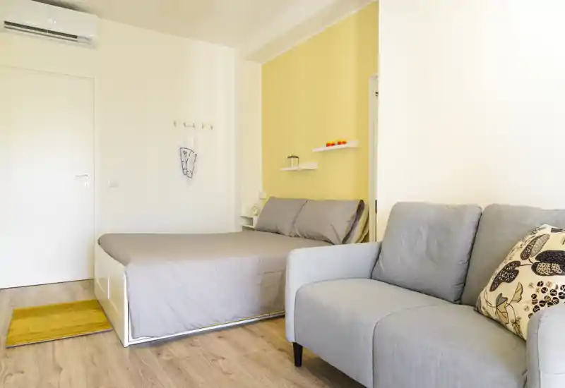 Camera da letto luminosa e ben arredata con letto matrimoniale, tessuti neutri e un piccolo divano in primo piano; parete d'accento gialla e mensole a muro creano un'atmosfera accogliente.