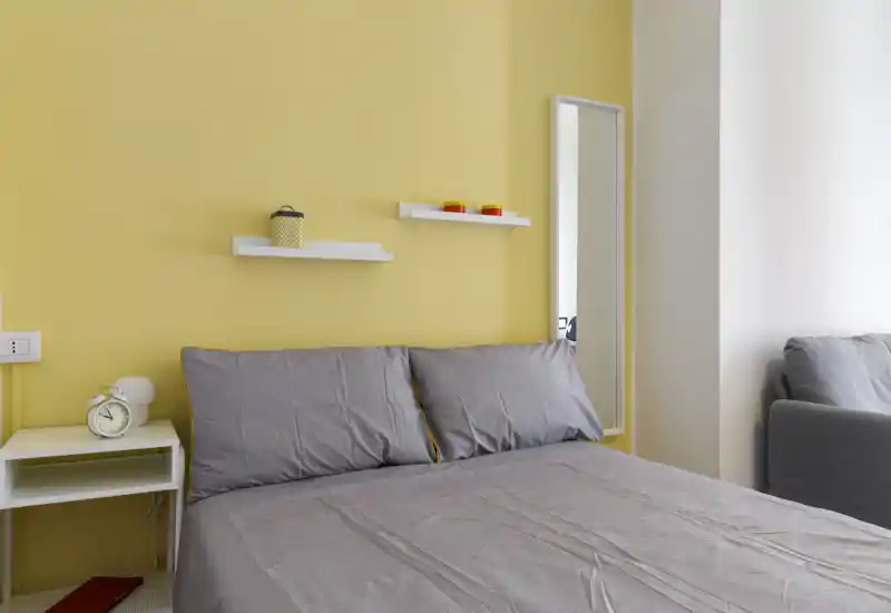 Camera da letto luminosa e moderna con un letto matrimoniale ben rifatto contro una parete gialla, mensole a muro e uno specchio a figura intera; biancheria grigia e comodino completano l'ambiente accogliente.