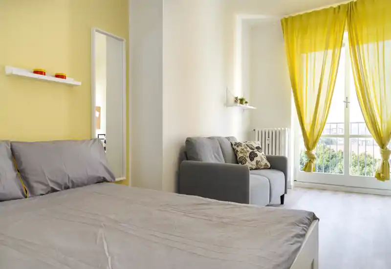 Camera da letto luminosa e accogliente con un ampio letto in lenzuola grigie, una poltrona e tende gialle che incorniciano la porta sul balcone.