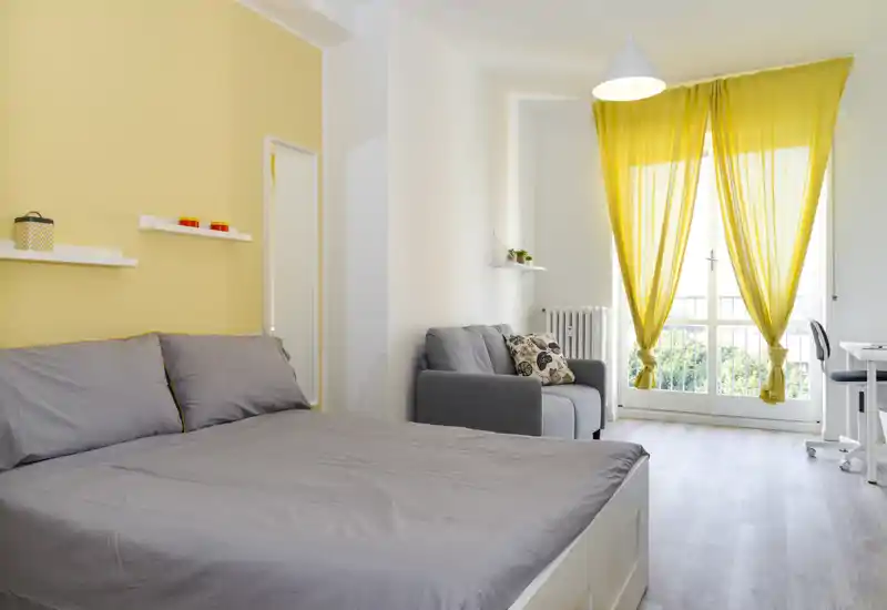 Camera da letto luminosa e moderna con un grande letto matrimoniale in primo piano, una poltrona grigia accogliente e una porta-finestra con tende gialle che illumina la stanza con luce naturale.