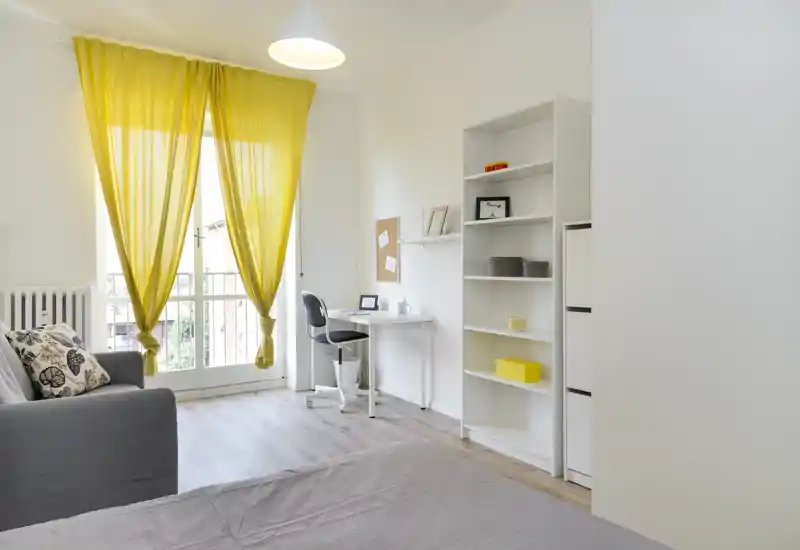 Camera da letto luminosa e moderna con letto in primo piano, una scrivania vicino alla grande finestra con tende gialle e scaffalatura aperta — elegante e ariosa.