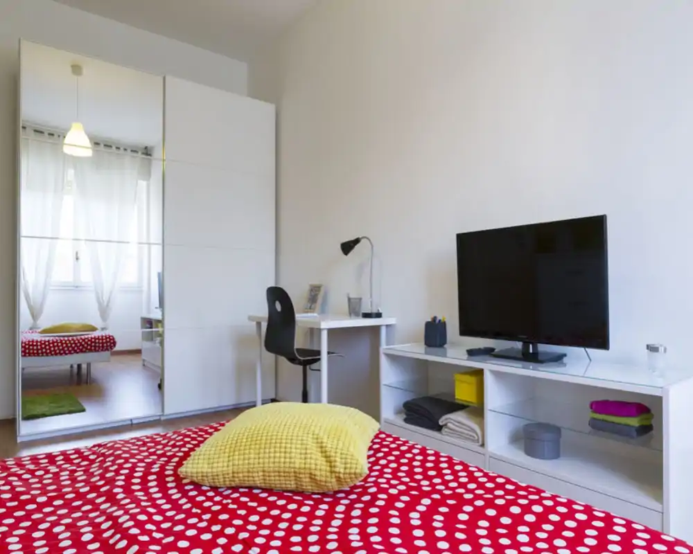 Dormitorio luminoso y bien decorado con funda de lunares roja, cojín amarillo y armario con espejo que refleja la luz. Escritorio y mueble TV completan este espacio acogedor y funcional.