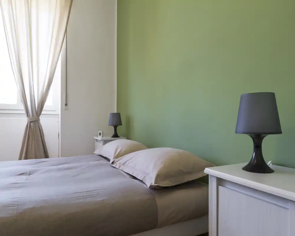 Dormitorio luminoso y minimalista con una cama doble bien hecha, mesillas y lámparas a juego frente a una pared verde suave. La luz natural entra por cortinas claras, creando un ambiente sereno y aireado, ideal para descansar.