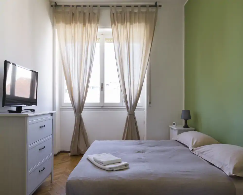 Dormitorio luminoso y ordenado con cama doble, ropa de cama suave, mesita con lámpara y luz natural de una gran ventana — ideal para estancias cómodas.