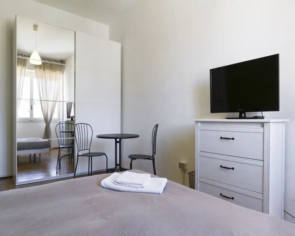 Dormitorio luminoso y minimalista con cama bien hecha, armario con espejo que refleja la luz natural, una pequeña mesa con dos sillas y cómoda con TV — espacio funcional y cómodo para estancias cortas.