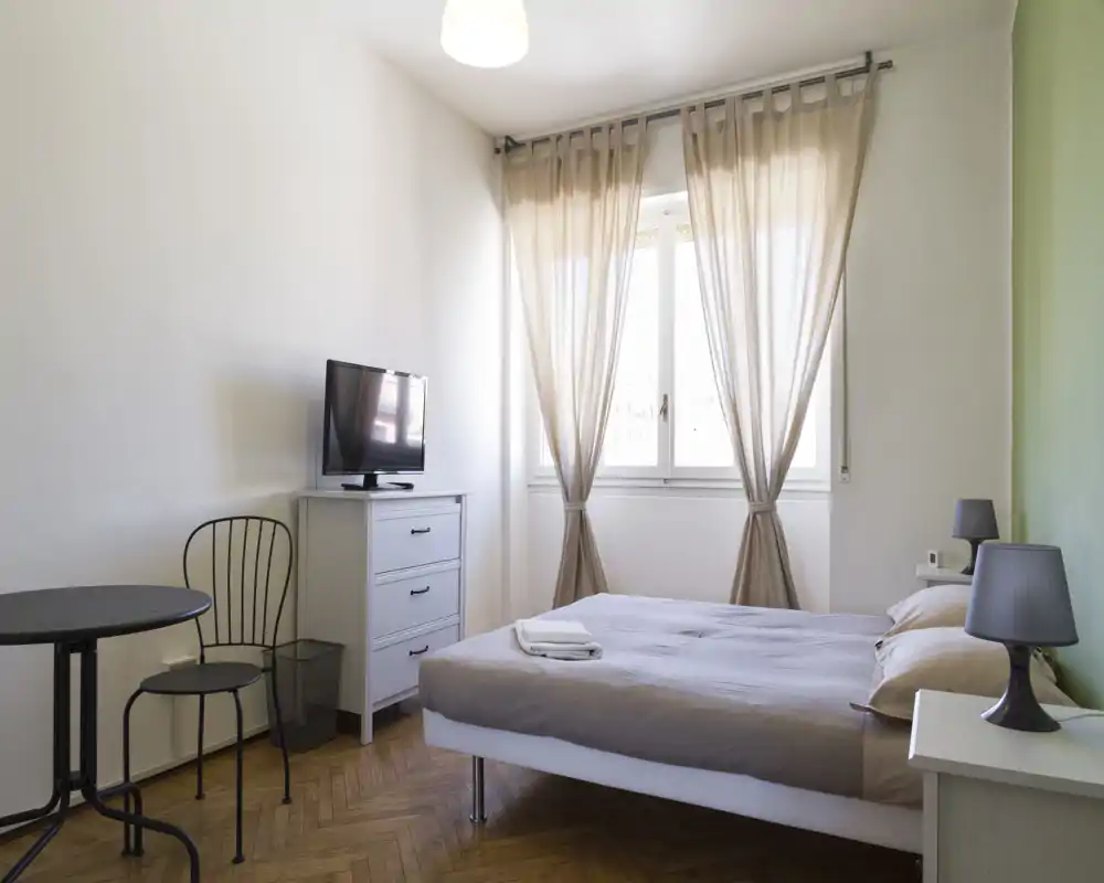 Dormitorio doble luminoso y bien ordenado con cama cómoda, cortinas suaves y lámparas de mesita. La habitación incluye una mesa pequeña con sillas, cómoda con TV y suelo de parquet cálido — ideal para alquileres de corta estancia.