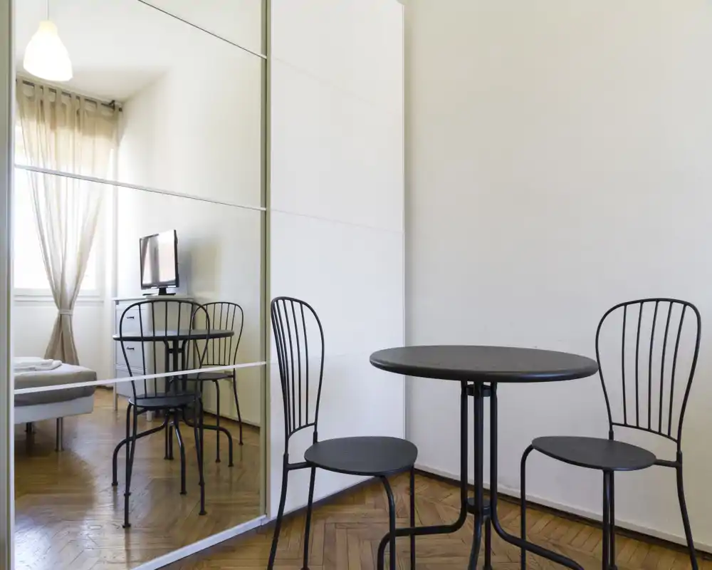 Zona de día luminosa y minimalista con una pequeña mesa redonda y sillas metálicas junto a un armario con espejo; luz natural desde la ventana y estilo moderno y sencillo.