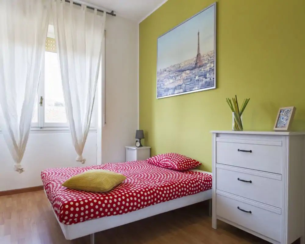 Dormitorio luminoso y ordenado con cama roja de lunares, suelo de madera clara y cómoda decorativa; gran ventana con luz natural suave.