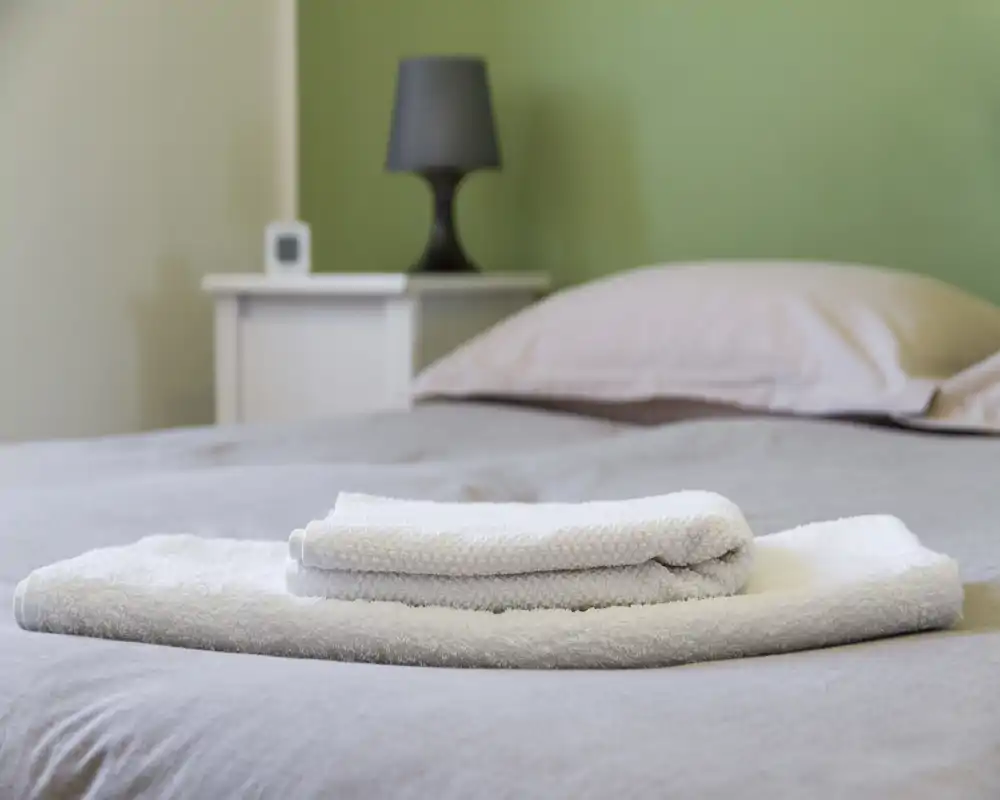 Dormitorio luminoso y ordenado con la cama en primer plano y toallas blancas dobladas; ropa de cama en tonos neutros, mesita y lámpara que aportan calidez.