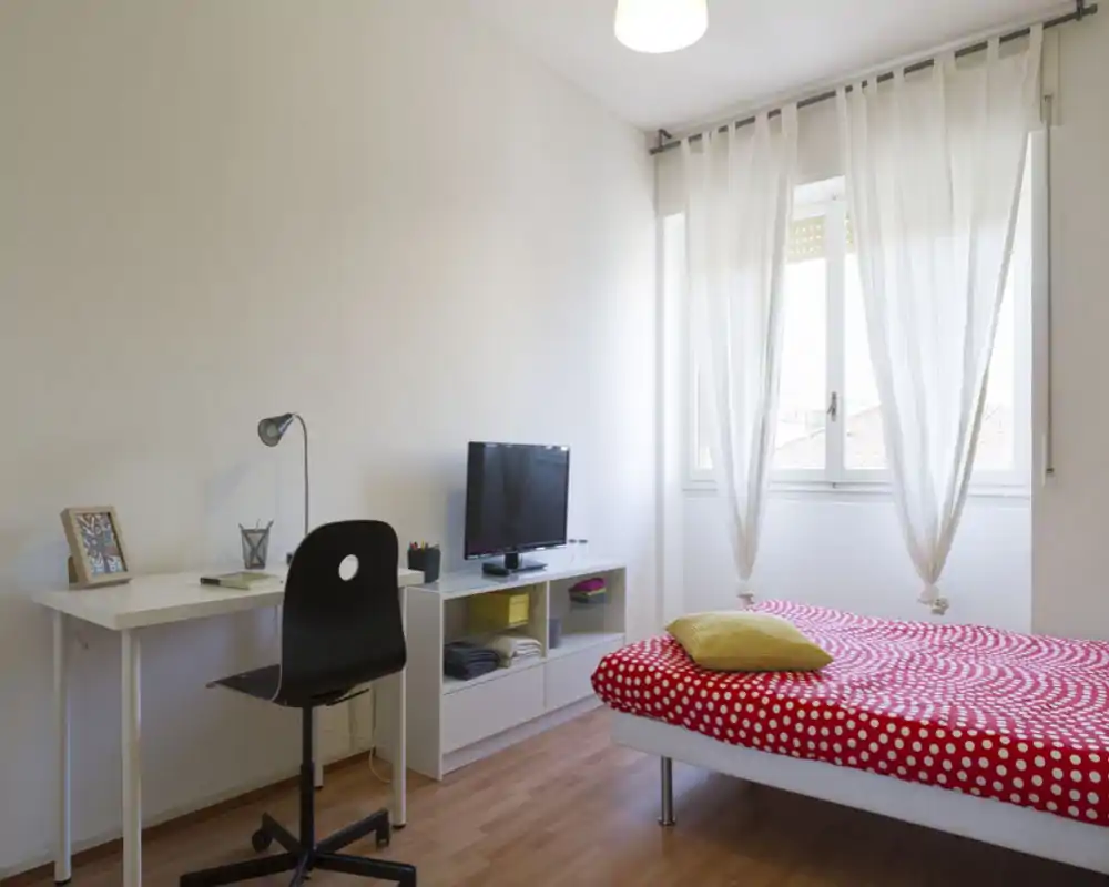 Dormitorio luminoso y moderno con funda roja de lunares, escritorio y mueble TV junto a una gran ventana que aporta luz natural suave.