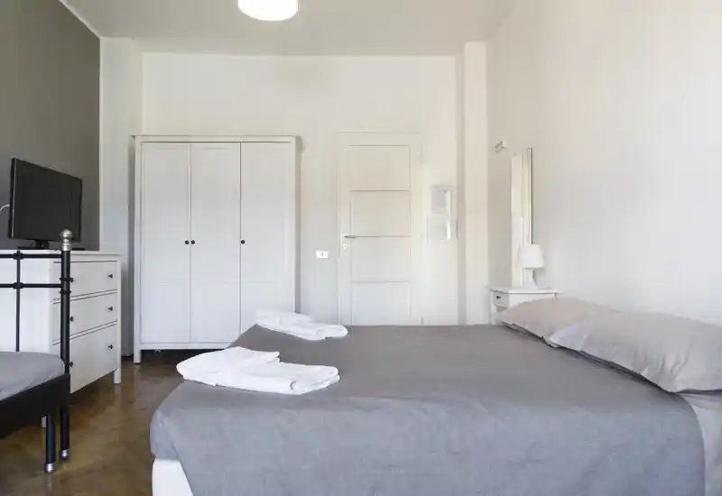 Bright, minimalist bedroom with a large double bed dressed in grey linens, white wardrobe and bedside table—clean, well-lit and ready for guests.