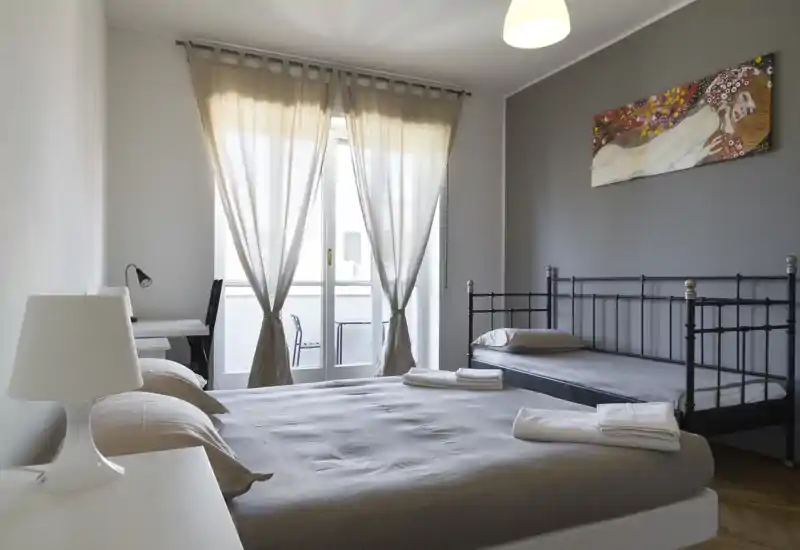 Bright, neatly styled bedroom with a double bed in the foreground and a single metal daybed against the wall; soft neutral linens, a desk by the window and balcony doors that fill the room with natural light.