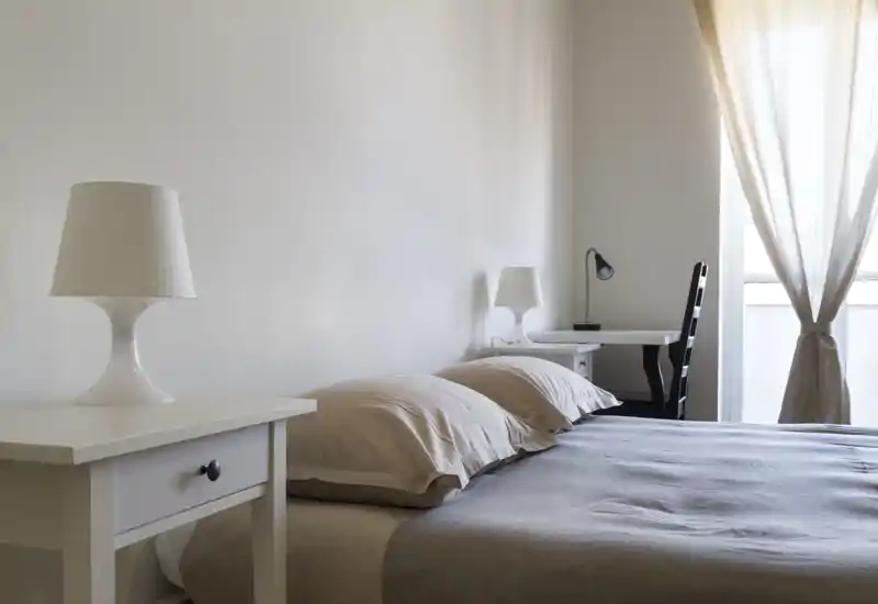 Minimalist, bright bedroom with a large bed, neutral linens, bedside tables and soft natural light from a curtained window.