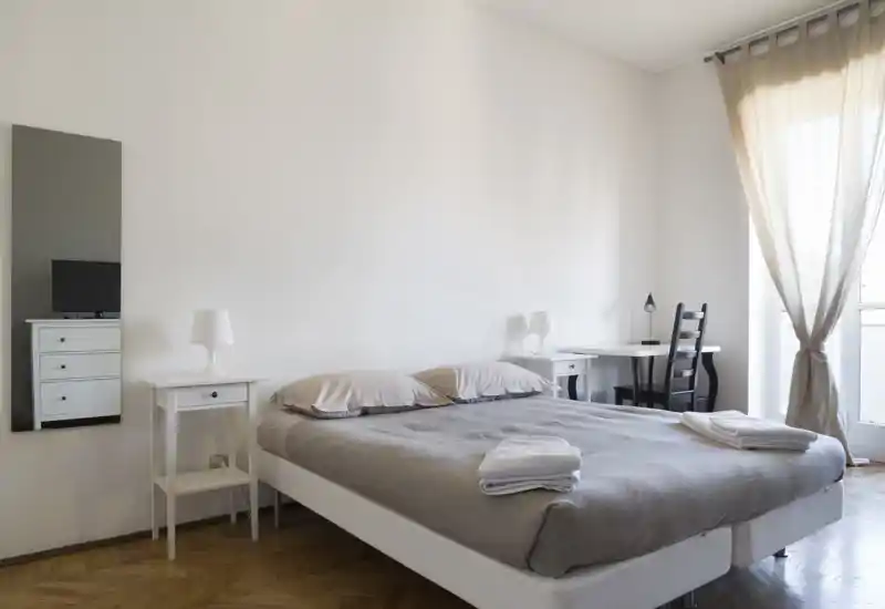 Bright, minimalist bedroom with a double bed, bedside tables and a small desk by the window — calm neutral tones and natural light.