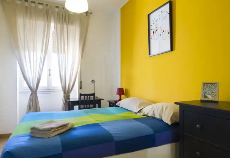 Bright, colorful bedroom with a double bed against a vivid yellow accent wall, natural light from a large window, bedside lamp and simple desk — a cozy, well-lit room ideal for rental listings.