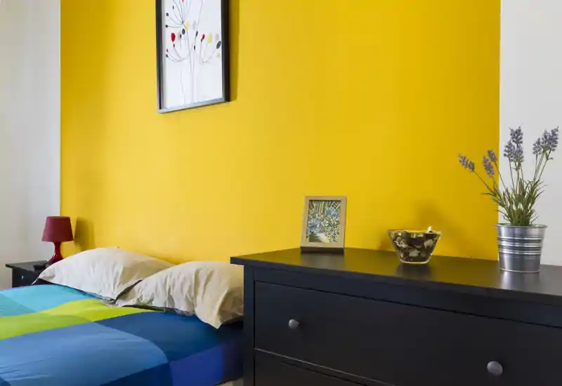 Dormitorio moderno y luminoso con cama colorida frente a una pared amarilla intensa, lámpara de noche y cómoda con elementos decorativos — ideal para anuncios.
