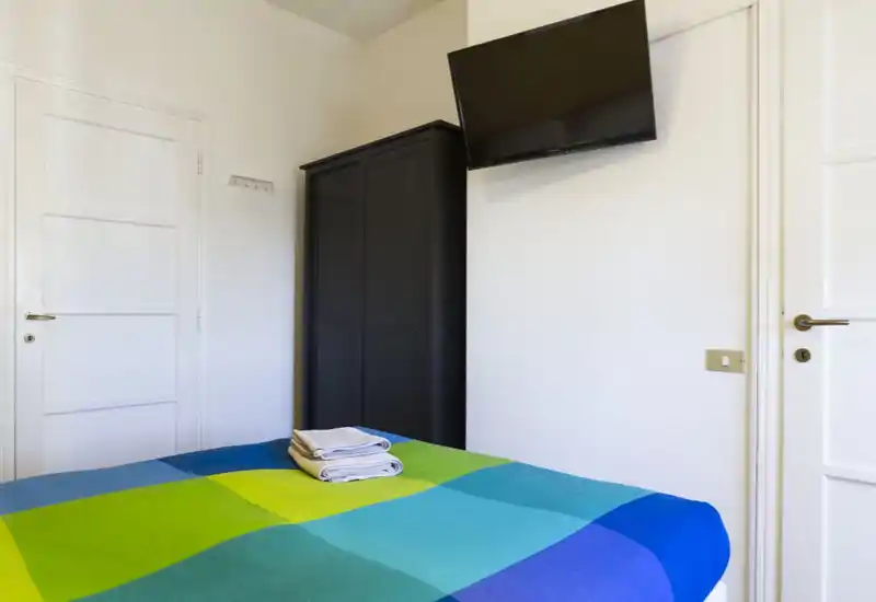 Dormitorio luminoso y sencillo con colcha colorida, toallas dobladas sobre la cama, armario y TV de pared — ideal para estancias cortas.
