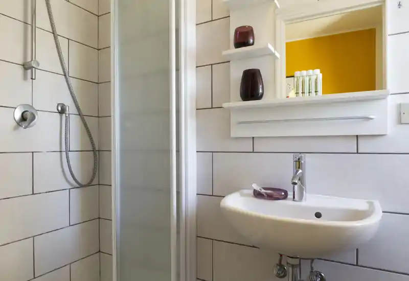 Baño compacto y moderno con lavabo mural, ducha de mano y mampara de vidrio esmerilado. Azulejos blancos y accesorios sencillos que crean un espacio limpio y funcional.