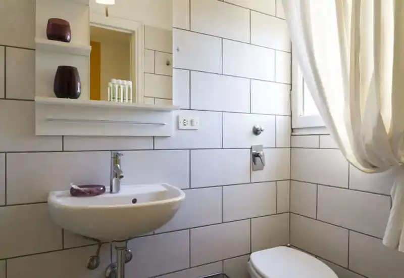 Baño compacto y luminoso con lavabo de pared, estante con espejo, azulejos y una ventana con cortina que aporta luz natural suave.