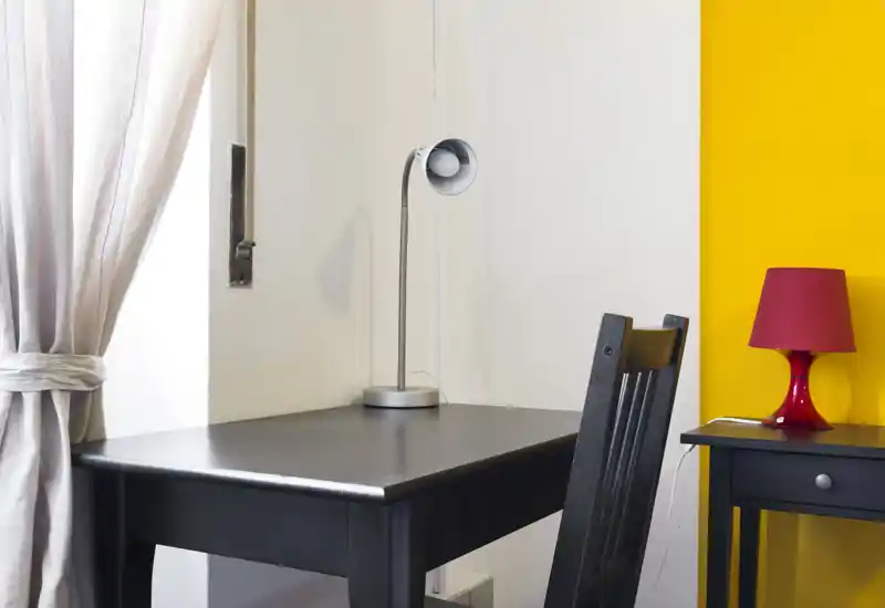 Rincón de estar/trabajo compacto con mesa y silla de madera oscura junto a una pared amarilla y luz natural de la ventana.