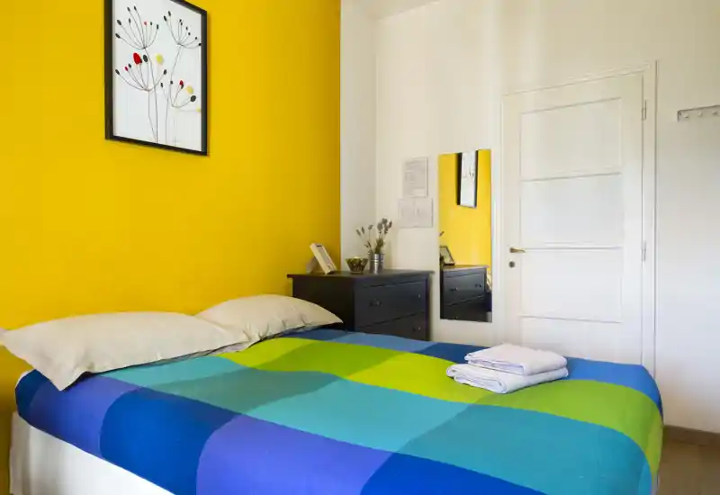 Bright, colorful bedroom with a double bed covered by a vivid blue-green check duvet, bedside chest of drawers and framed wall art on a yellow accent wall.