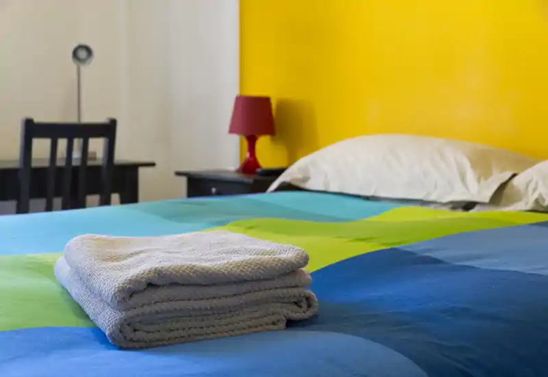 Dormitorio luminoso y colorido con cama bien hecha, toallas dobladas sobre la colcha y mesita con lámpara — pared amarilla vibrante que añade carácter.