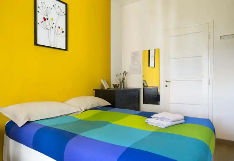 Dormitorio luminoso y colorido con cama doble vestida con ropa de cama azul y verde, dos almohadas y toallas dobladas. Pared de acento amarilla, cuadro y cómoda negra con objetos decorativos que hacen el espacio acogedor.