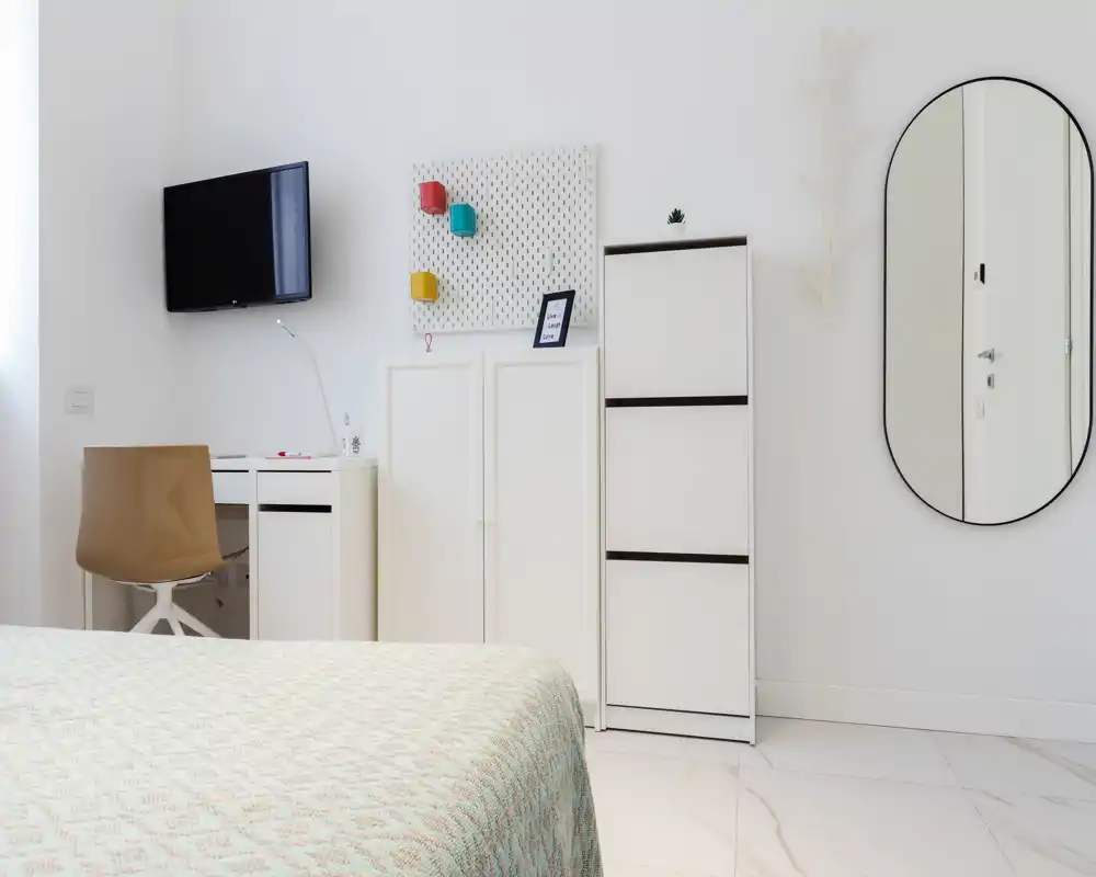 Bright, minimal bedroom featuring a neatly made bed in the foreground, white storage units, a wall-mounted TV and an oval mirror. Clean, modern styling and abundant natural light make the space feel fresh and airy.