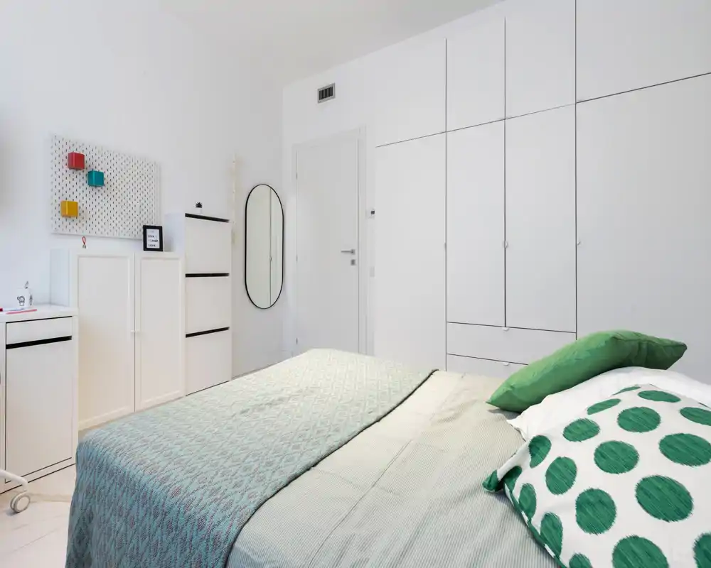 Bright, modern bedroom with a neatly made double bed, white built-in wardrobes and minimalist decor featuring green accent pillows and patterned bedding.