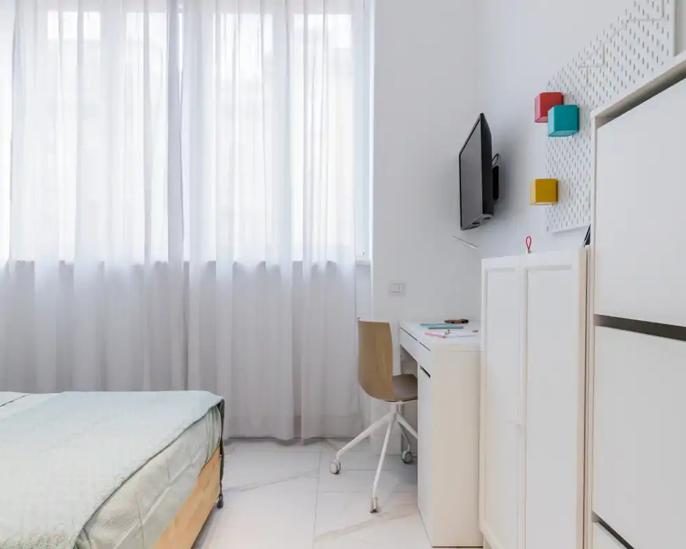Bright, minimal bedroom with a partially visible bed, a small desk and chair by the window, and neat storage units — ideal for a single occupant or short stays.
