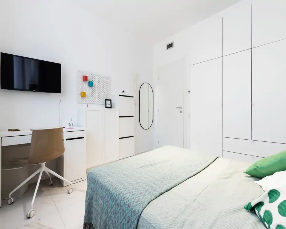 Bright, modern bedroom with a neatly made bed, built-in white wardrobes and a small desk. Clean minimalist styling with soft textiles and pleasant natural light.