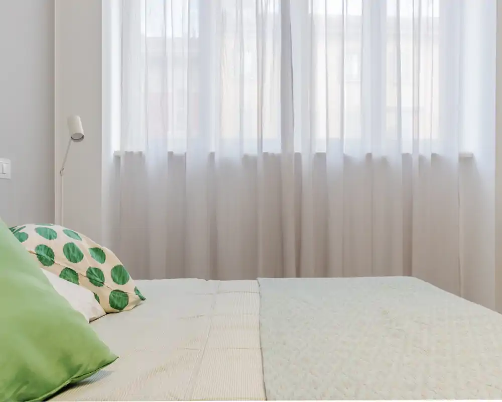 Bright, softly styled bedroom view with a neatly made bed, patterned cushions and light filtering through sheer curtains for a calm atmosphere.