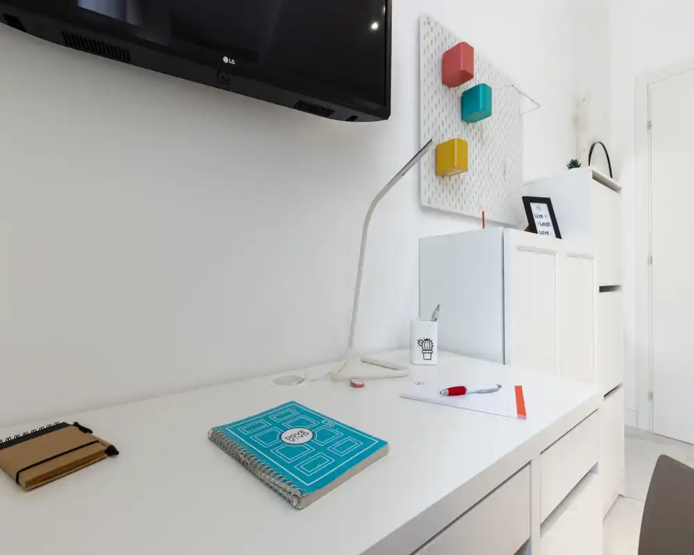 Bright, minimal living area corner with a white desk, wall-mounted TV, and decorative storage elements — ideal for remote work or relaxing.