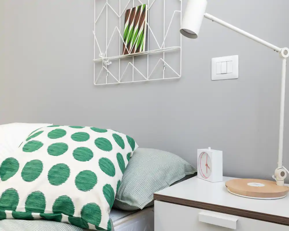 Close-up of a styled bedroom corner showing pillows, mattress edge and a bedside table with lamp and clock; clean, modern neutral tones with a playful green polka-dot cushion.